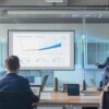 a sleek, modern office interior features an animated presentation on a large screen, highlighting the stark differences in energy costs associated with various roofing materials, with focused professionals engaging in discussion around a stylish conference table.